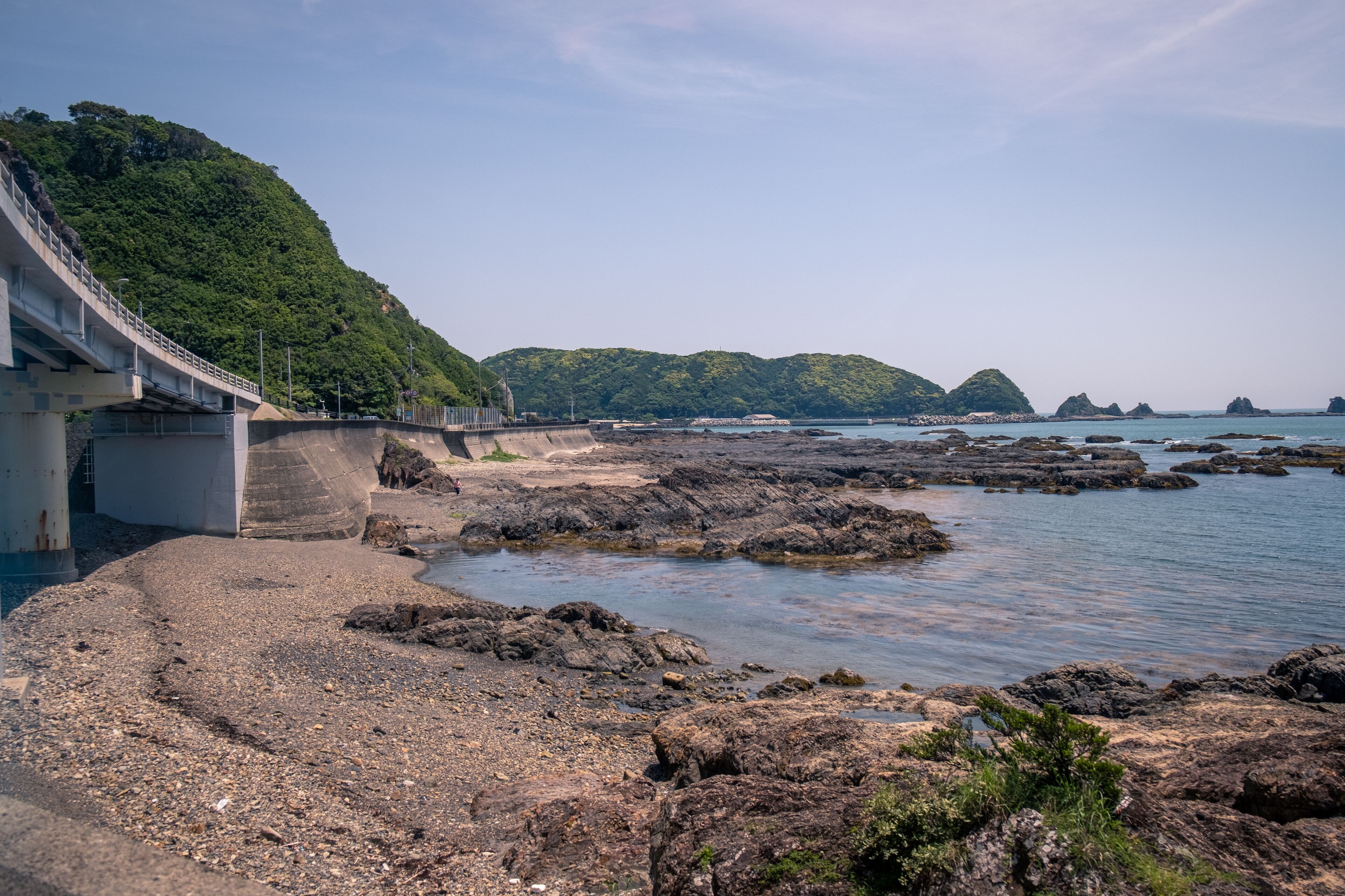 A coastal landscape with rocky shores, a bridge, and lush green hills in the background.