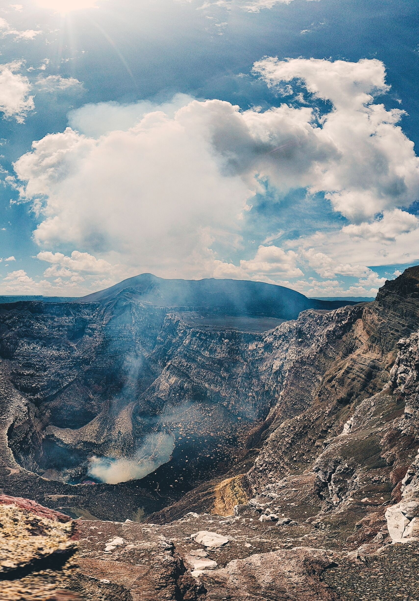 A volcanic crater with smoke rising, surrounded by rugged terrain and a cloudy sky.