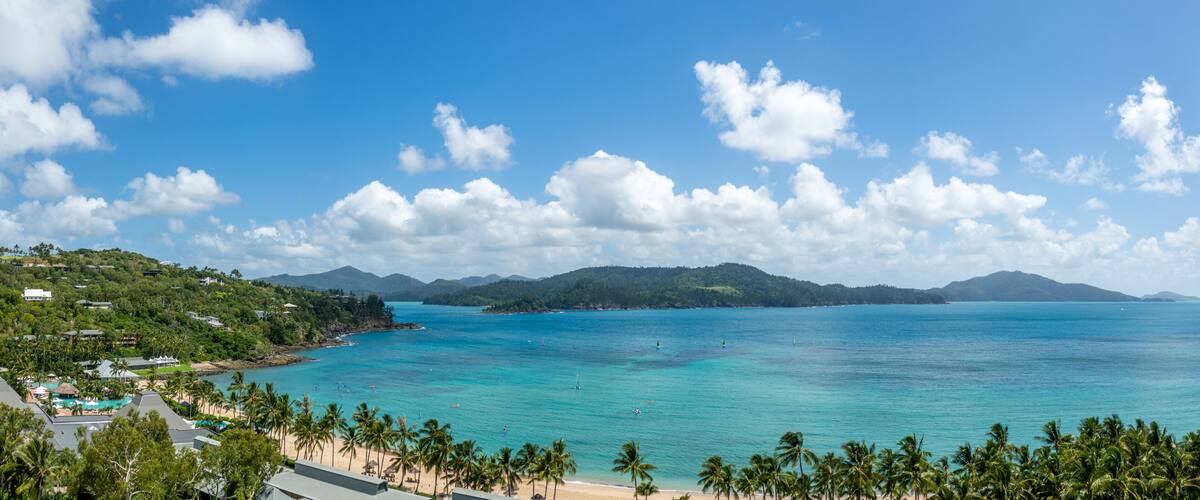 A tropical beach with clear turquoise waters, palm trees, and distant mountains under a blue sky with scattered clouds.