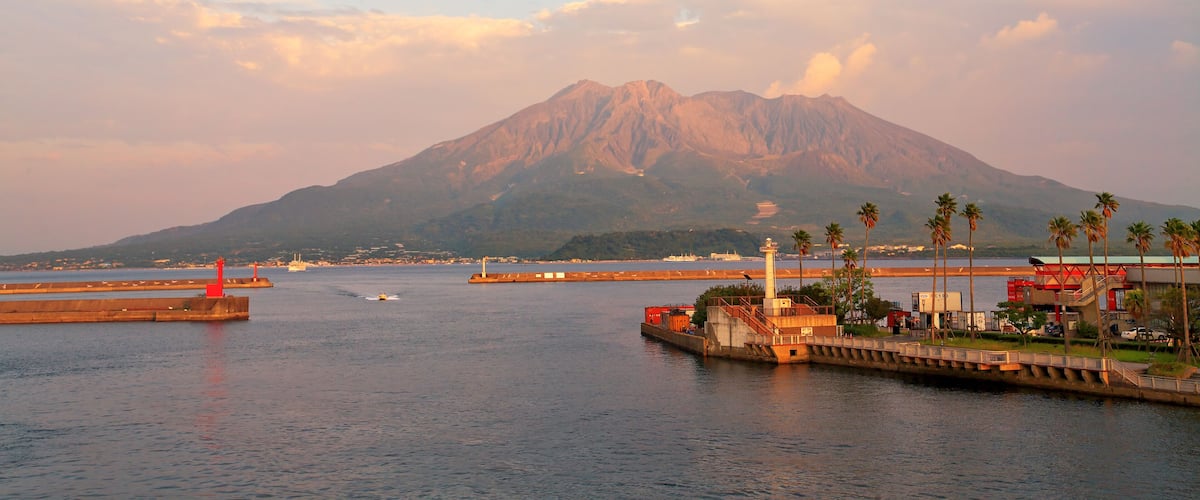 Escena costera con una montaña al fondo, un puerto con barcos y un faro rojo.