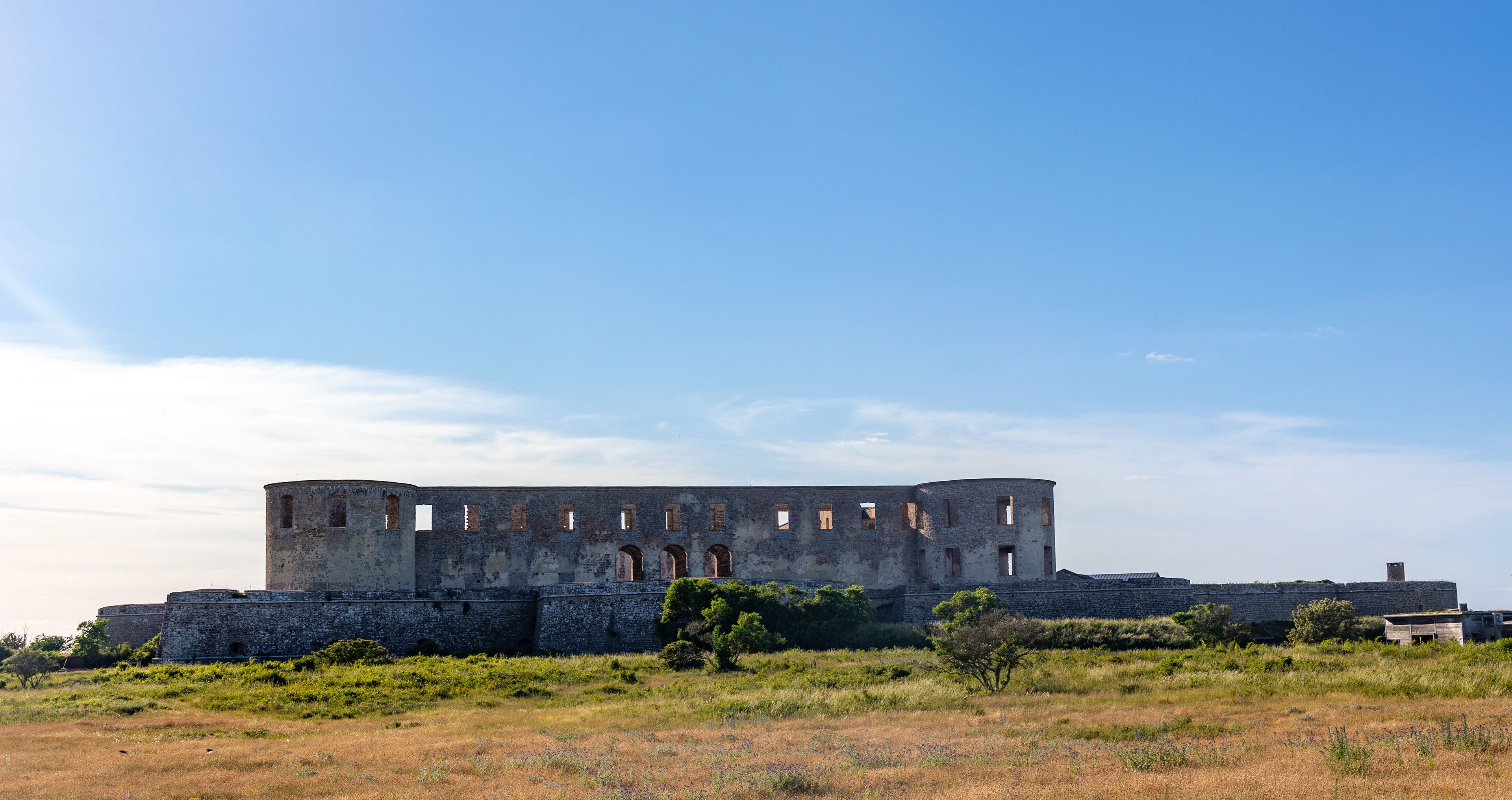 An ancient stone fortress with arched windows and a circular tower, surrounded by grassy fields and sparse vegetation.