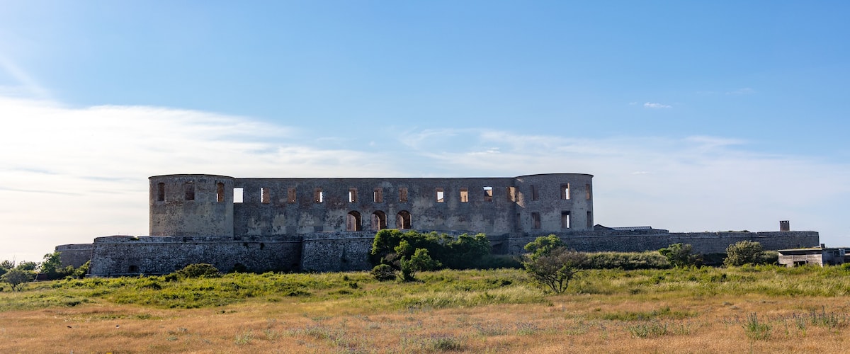 An ancient stone fortress with arched windows and a circular tower, surrounded by grassy fields and sparse vegetation.