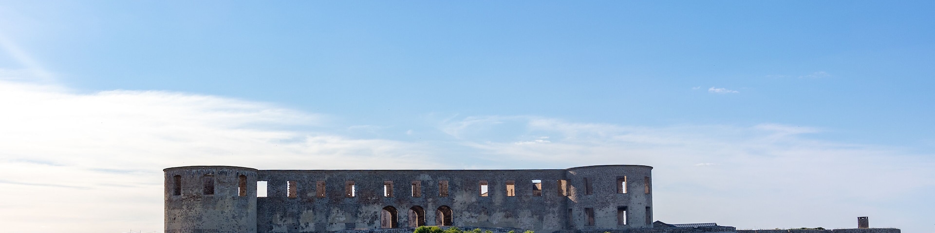 An ancient stone fortress with arched windows and a circular tower, surrounded by grassy fields and sparse vegetation.
