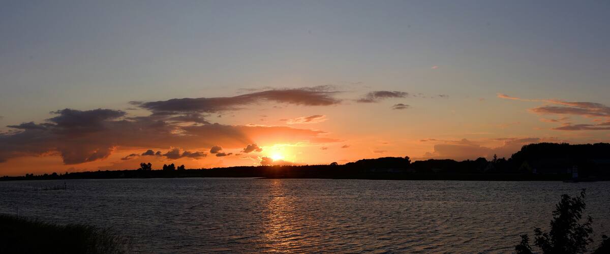A sunset over a calm lake with silhouettes of trees and buildings on the horizon.
