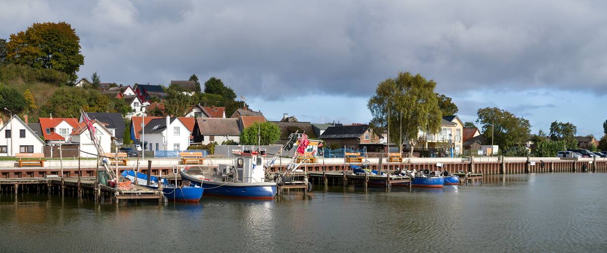 A harbor with boats docked, a wooden pier, and houses with red roofs in the background.