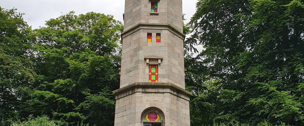 A stone tower with a staircase leading up to a door, surrounded by greenery and trees.