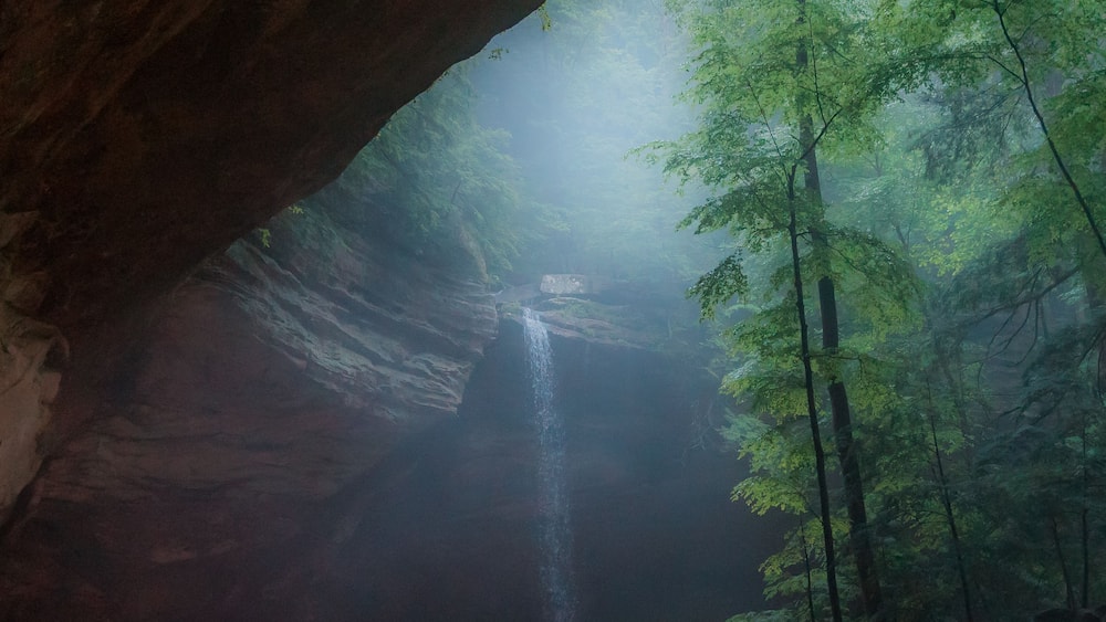 Une ouverture menant à une grotte, à l’intérieur de laquelle se trouve une cascade, le tout étant entouré de forêt.