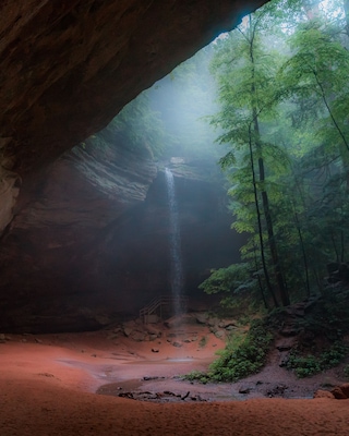 A cave opening with a waterfall inside, surrounded by forest.
