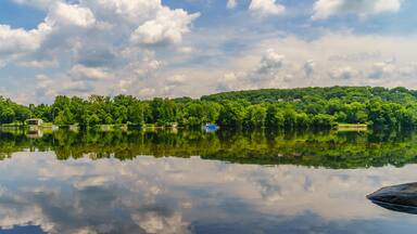 A calm lake with reflections of trees and clouds, surrounded by a forested hillside.
