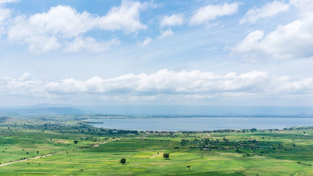 A panoramic view of a coastal landscape with rolling green fields, a large body of water, and distant mountains under a partly cloudy sky.