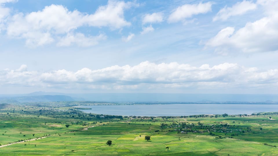 A panoramic view of a coastal landscape with rolling green fields, a large body of water, and distant mountains under a partly cloudy sky.