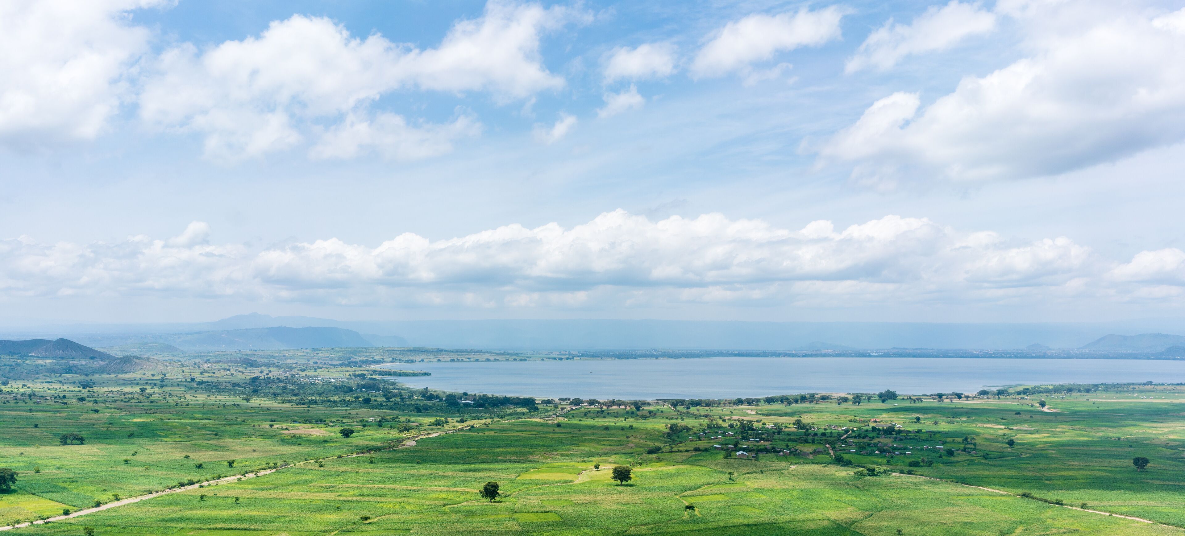A panoramic view of a coastal landscape with rolling green fields, a large body of water, and distant mountains under a partly cloudy sky.