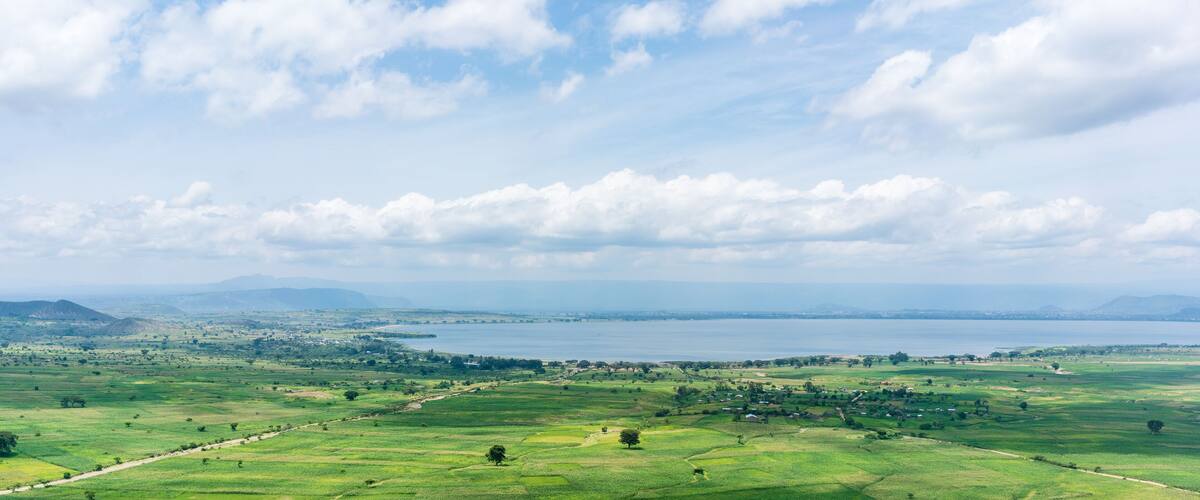 A panoramic view of a coastal landscape with rolling green fields, a large body of water, and distant mountains under a partly cloudy sky.