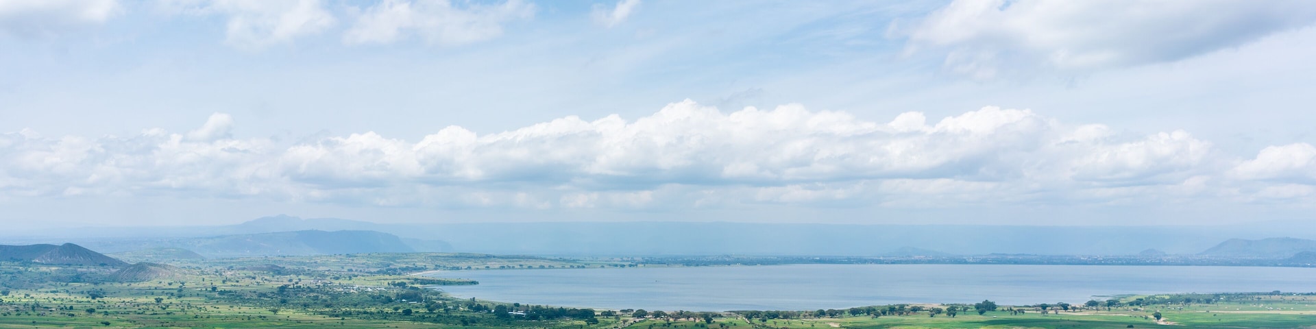 A panoramic view of a coastal landscape with rolling green fields, a large body of water, and distant mountains under a partly cloudy sky.