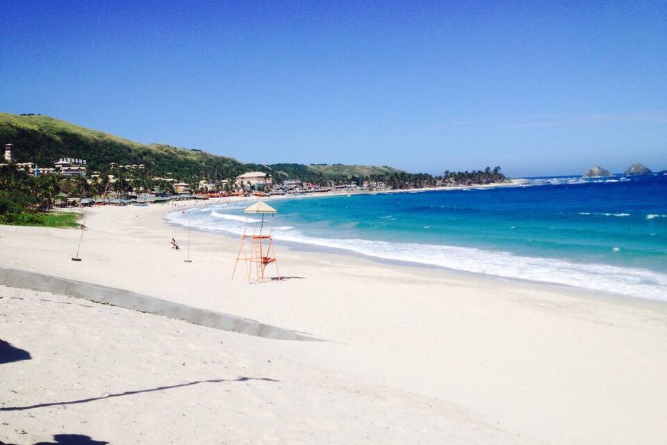 Uma praia com águas turquesa cristalinas, uma torre de salva-vidas e um horizonte distante com edifícios e colinas.
