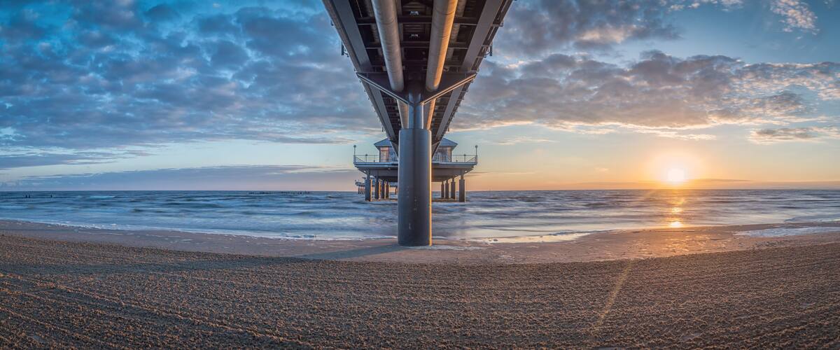 A beach with a pier extending into the ocean, a lifeguard tower, and the sun setting over the water.