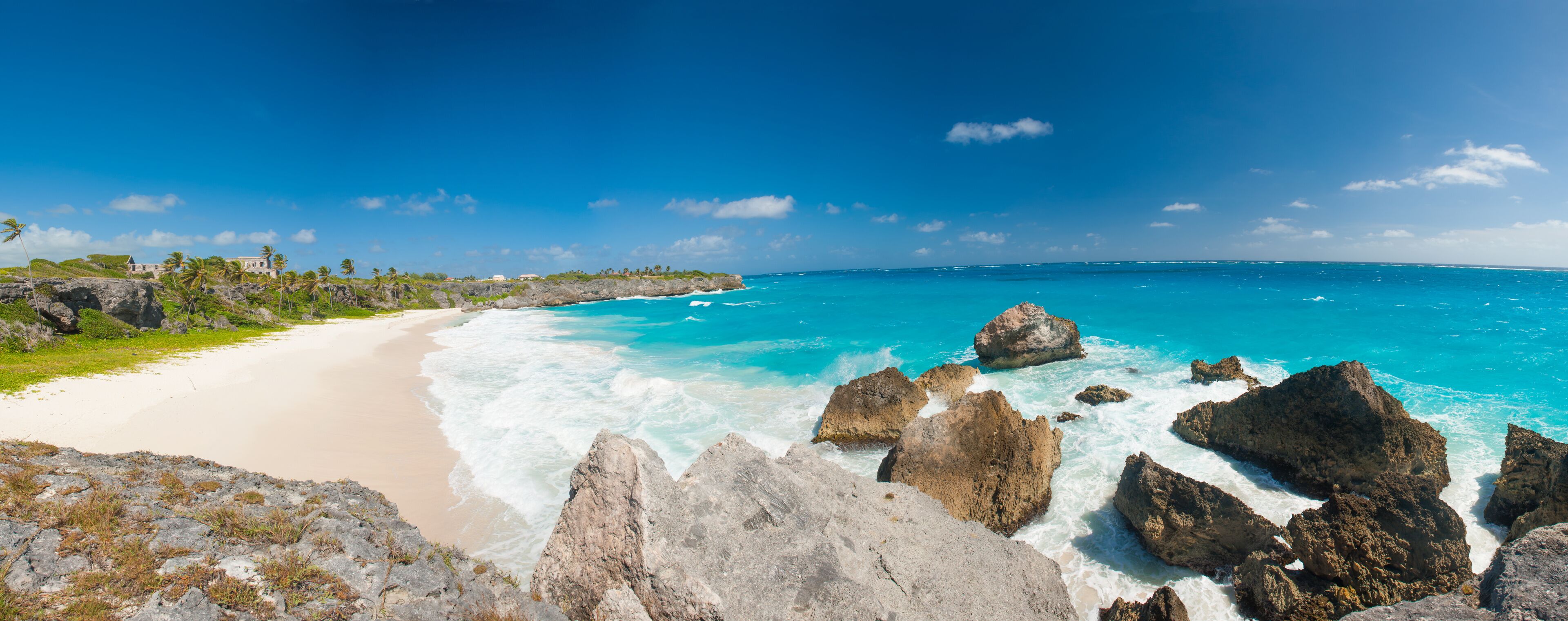 A beach with clear turquoise waters, rocky outcrops, and a distant building on a grassy hill.