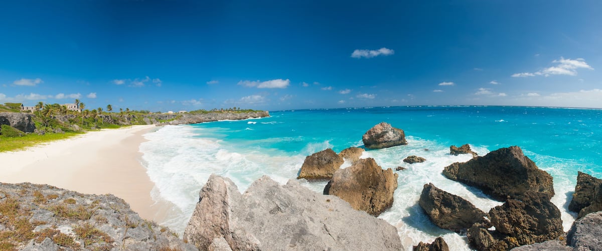 A beach with clear turquoise waters, rocky outcrops, and a distant building on a grassy hill.