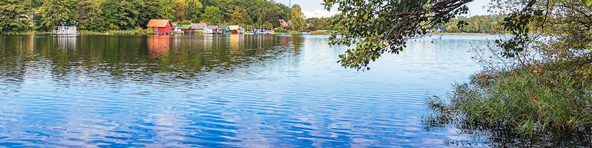 A calm lake with reflections, a red-roofed building, and a forested hill in the background.