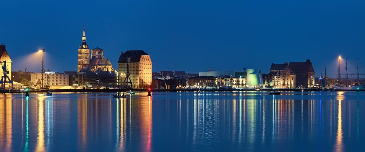 A city skyline at dusk with illuminated buildings and a calm river reflecting the lights.
