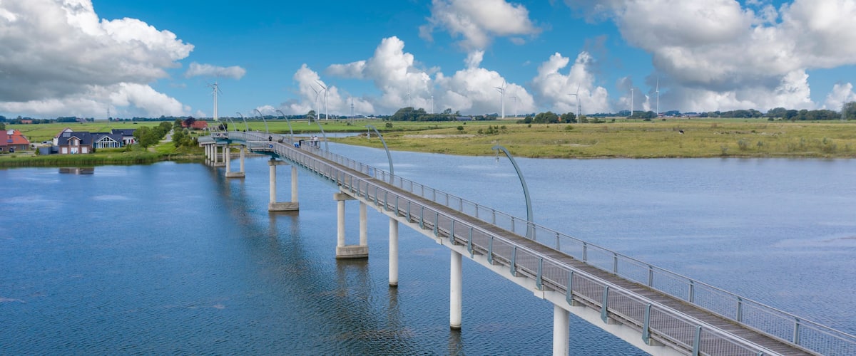 A long bridge over a calm river with a clear sky and wind turbines in the background.