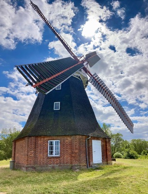 Un moulin à vent traditionnel, doté d’un toit incliné et d’une grande voile, se dressant sur un ciel partiellement nuageux.