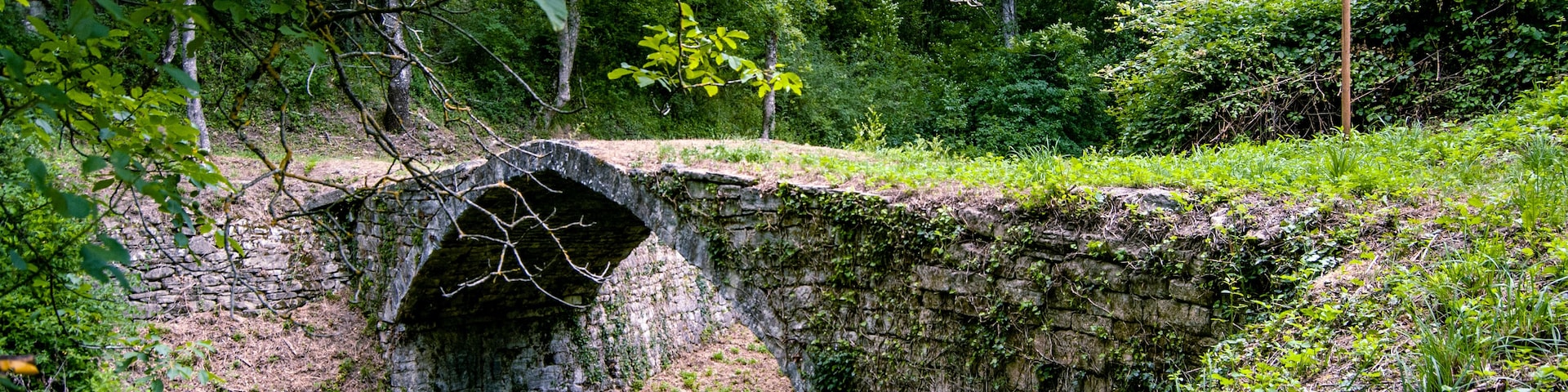 Eine steinerne Brücke überspannt ein schmaler Wasserlauf, umgeben von üppigem Grün und Bäumen.