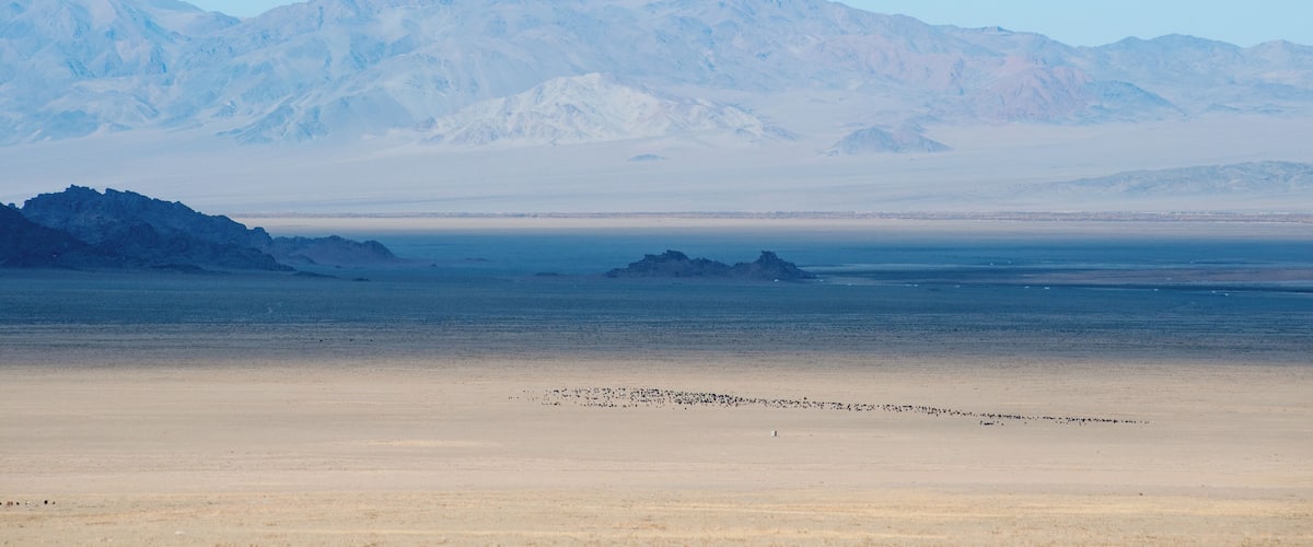 A vast desert landscape with a flat sandy foreground, a calm body of water, and distant snow-capped mountains under a clear sky.