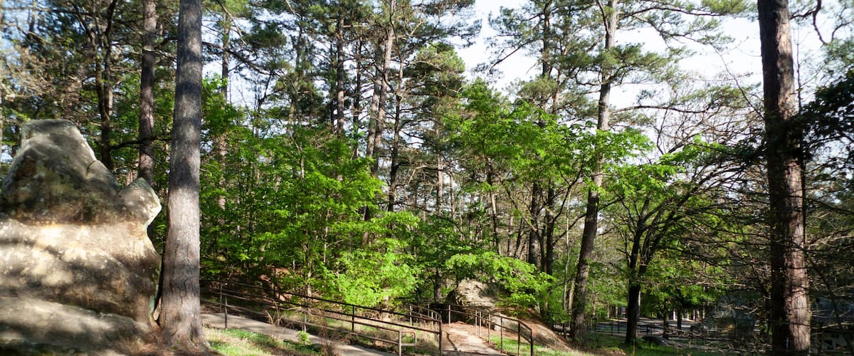 A forested area with a dirt path, metal railing, and a large rock formation.