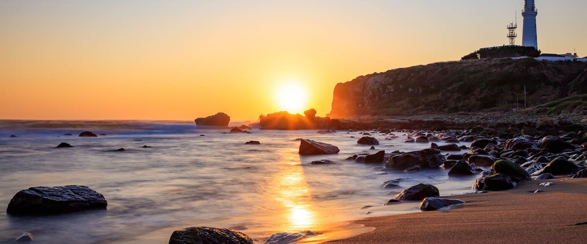 A sunset over a rocky beach with a lighthouse.