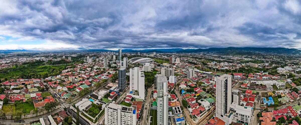 A cityscape with a mix of residential and commercial buildings, a prominent stadium, and a mountainous backdrop under a cloudy sky.
