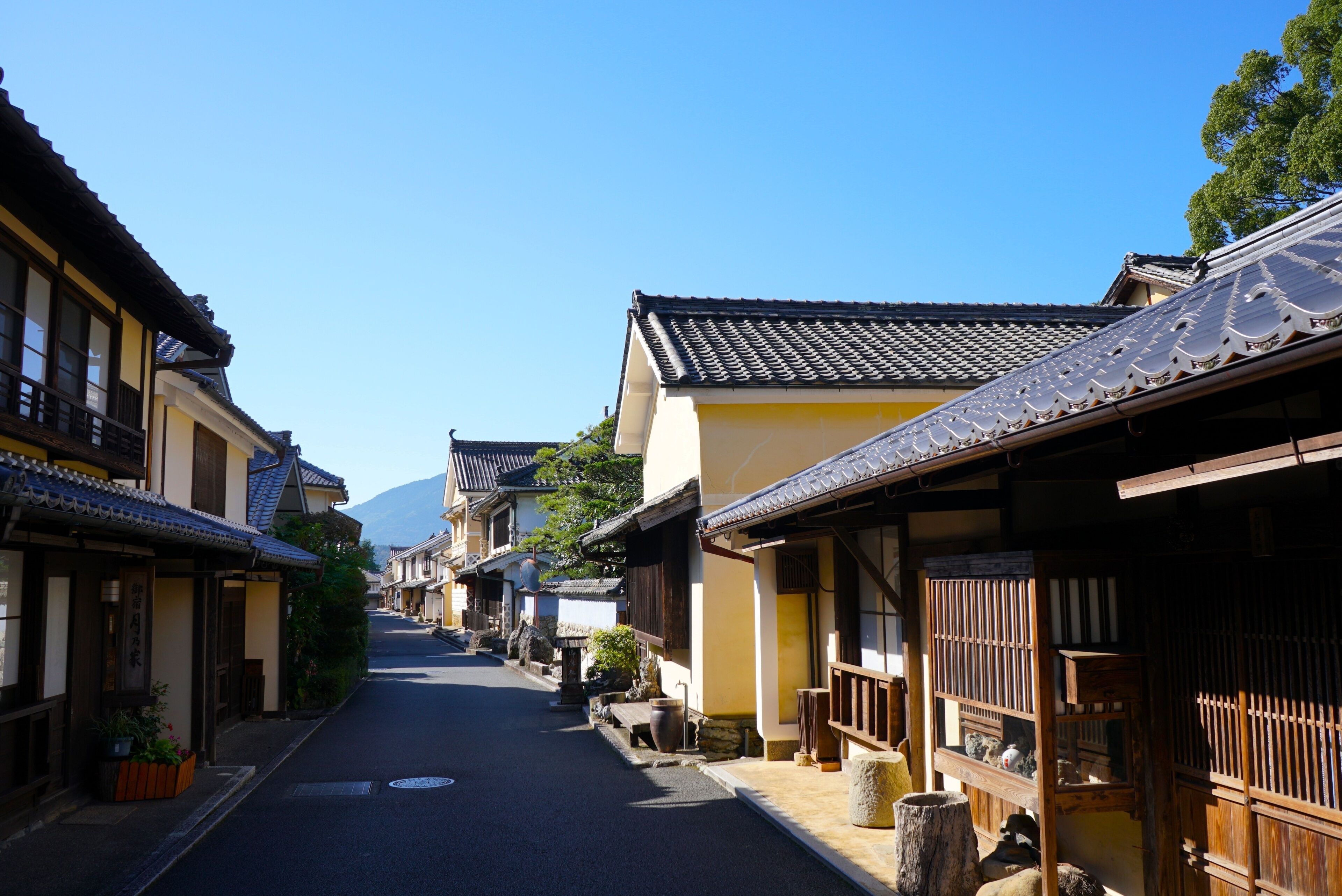 A narrow street lined with traditional Japanese buildings, a clear blue sky, and a mountain in the distance.