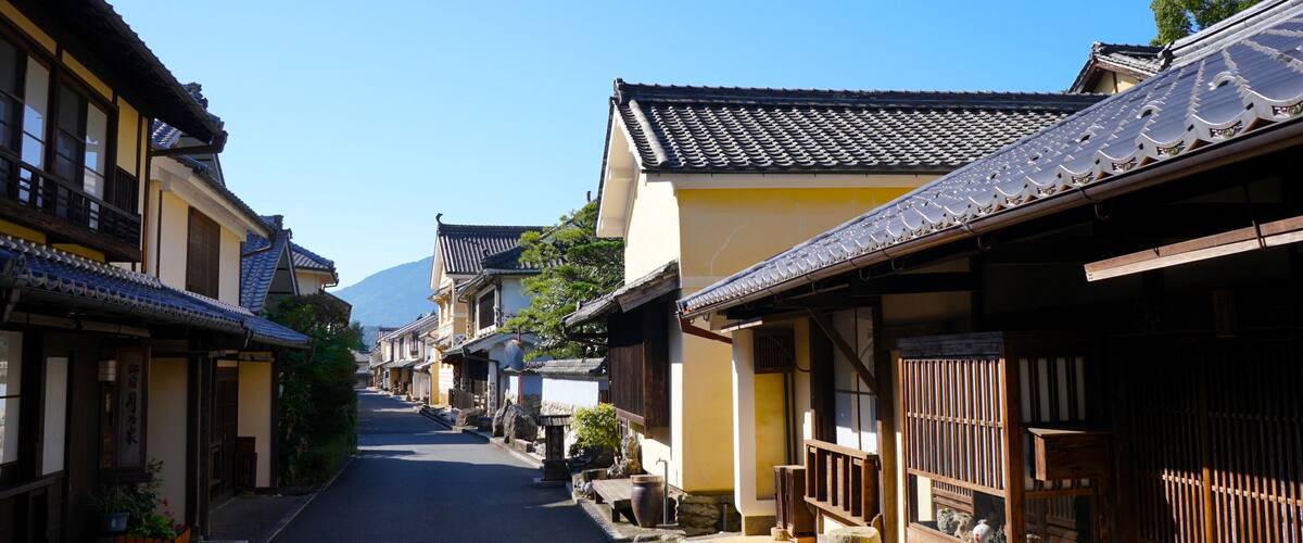 A narrow street lined with traditional Japanese buildings, a clear blue sky, and a mountain in the distance.