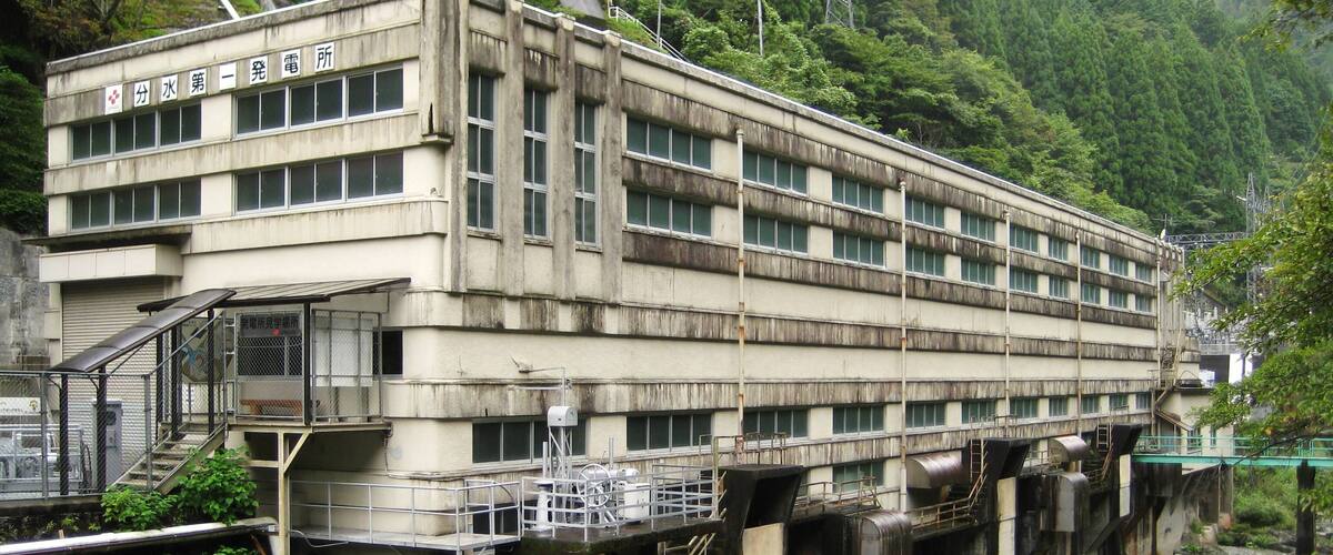 A large industrial building with multiple windows, elevated on stilts over a river, surrounded by lush greenery.