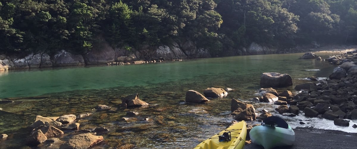 A calm river with clear water, surrounded by rocky shores and dense forested hills.
