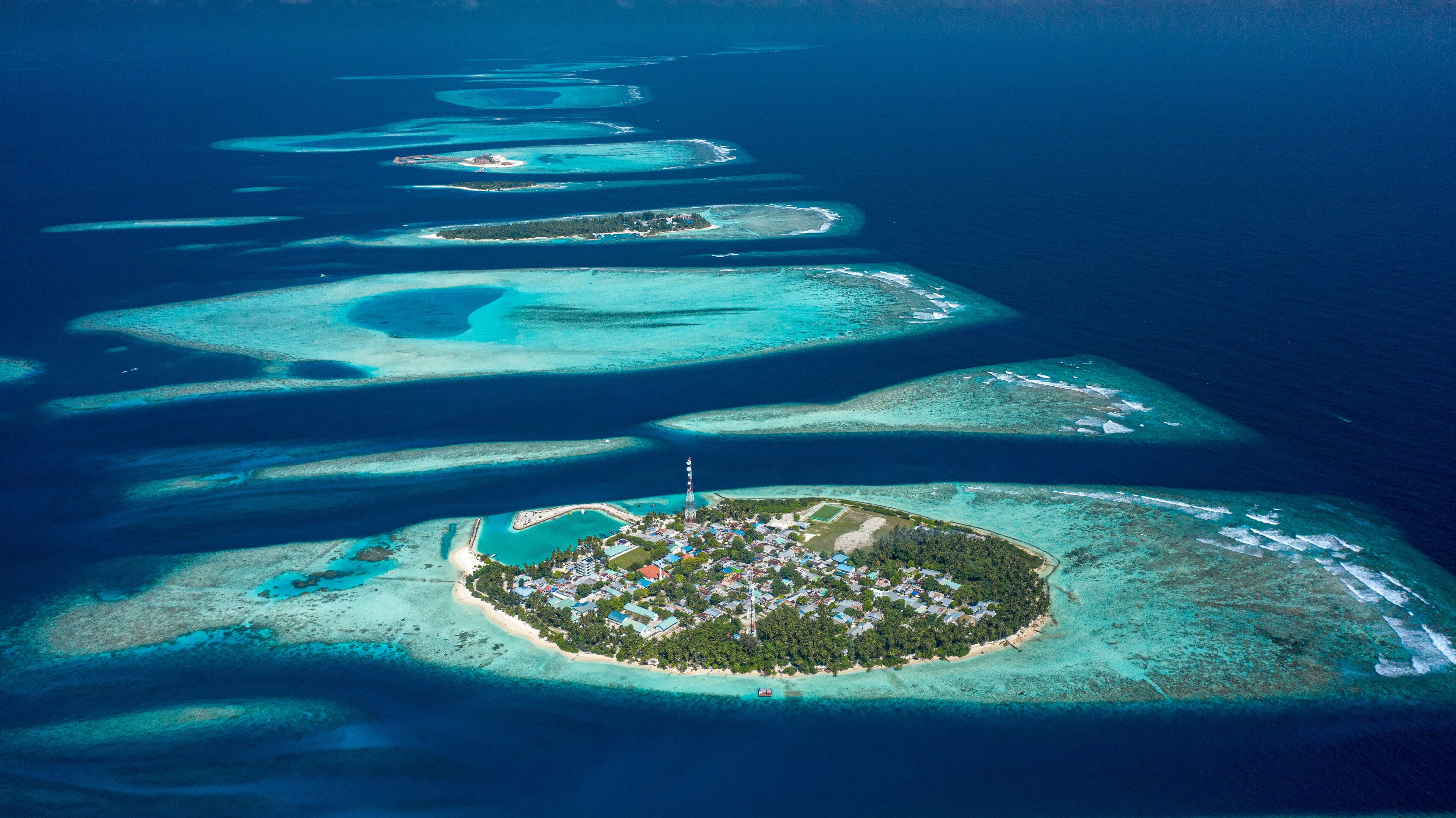 An aerial view of a chain of turquoise atolls with a central island featuring a dense settlement and a prominent lighthouse.