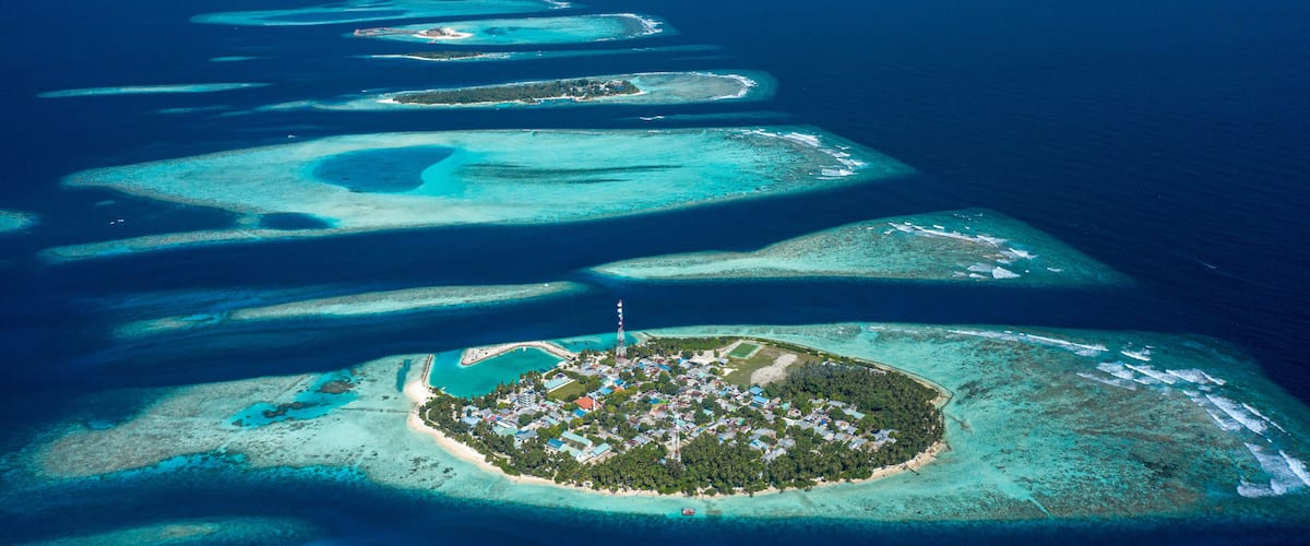 An aerial view of a chain of turquoise atolls with a central island featuring a dense settlement and a prominent lighthouse.