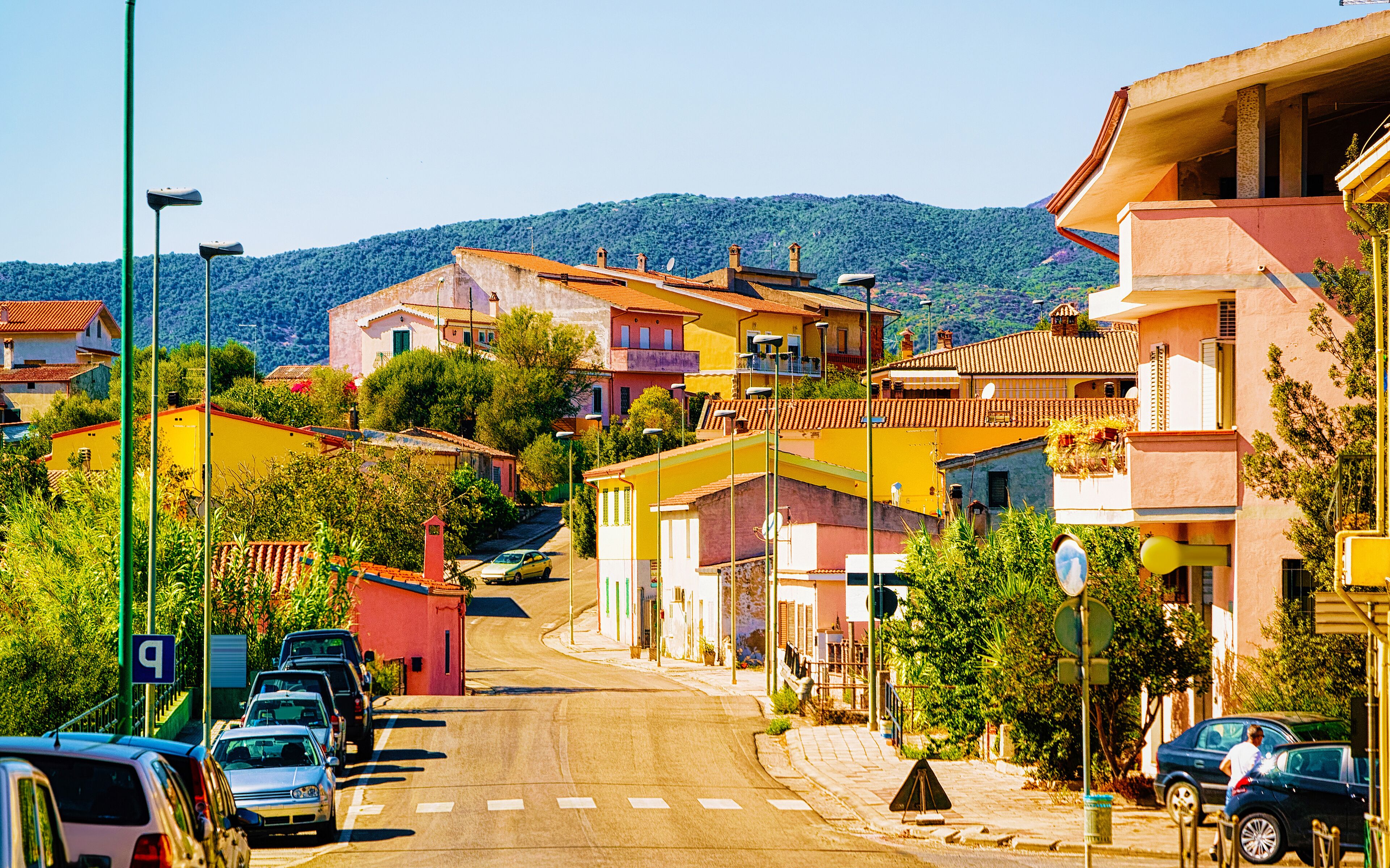 A street lined with colorful buildings, parked cars, and a hilly backdrop.
