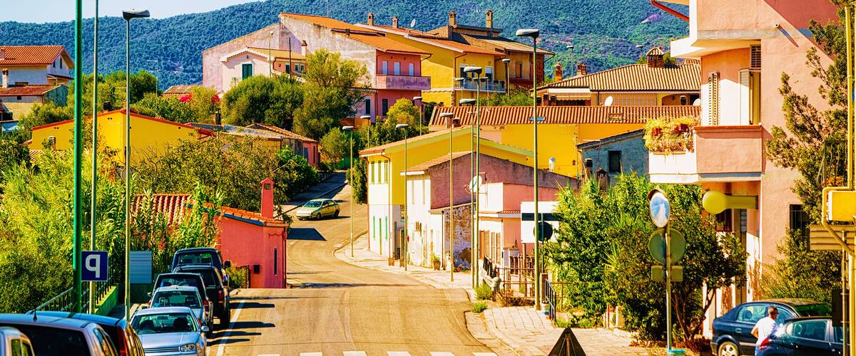 A street lined with colorful buildings, parked cars, and a hilly backdrop.