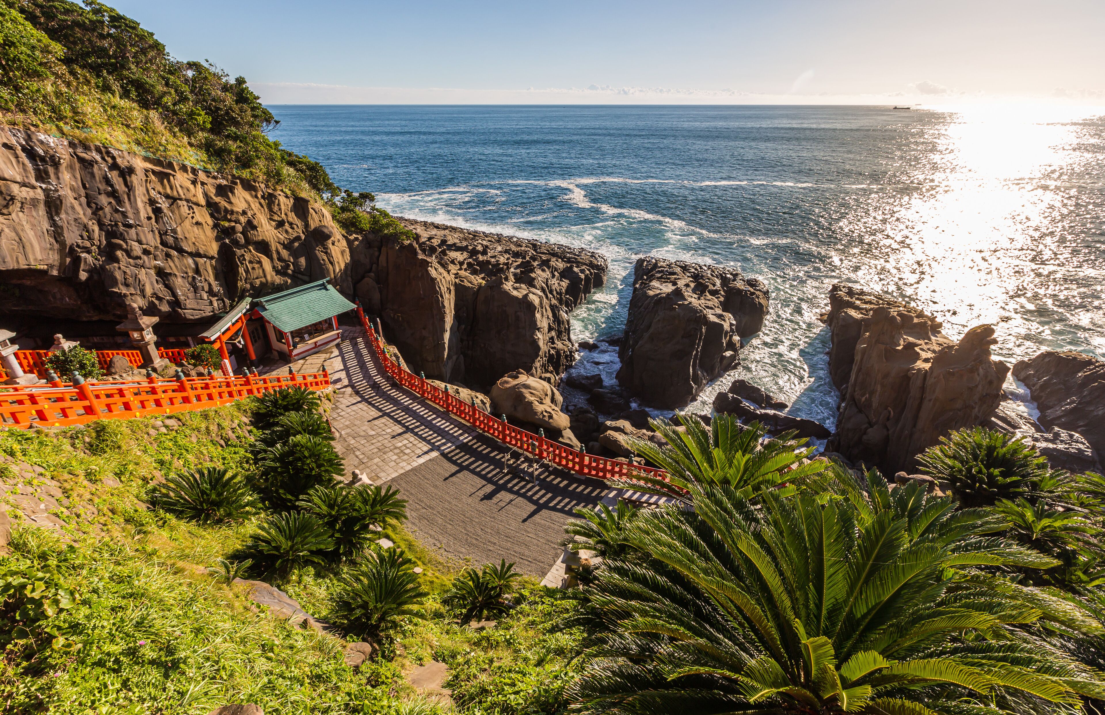 A coastal landscape with rocky cliffs, a temple with a green roof, and palm trees.