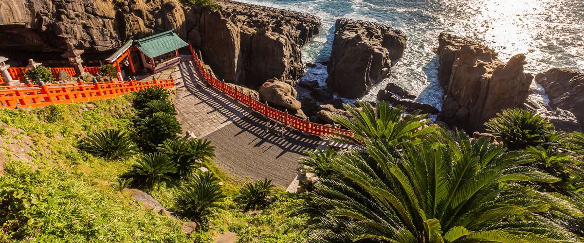 A coastal landscape with rocky cliffs, a temple with a green roof, and palm trees.