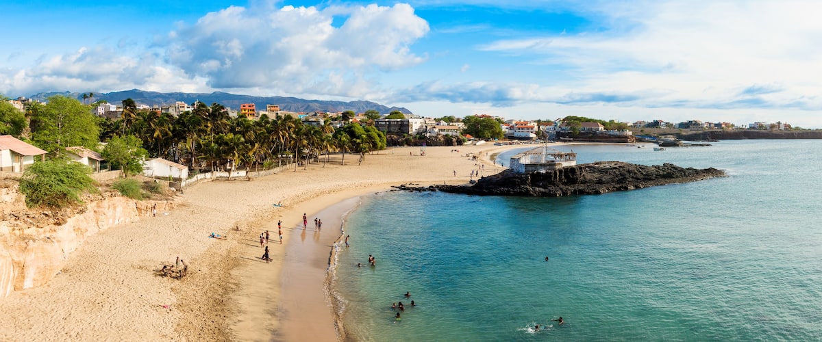 A coastal town with a sandy beach, clear blue waters, and a rocky outcrop.