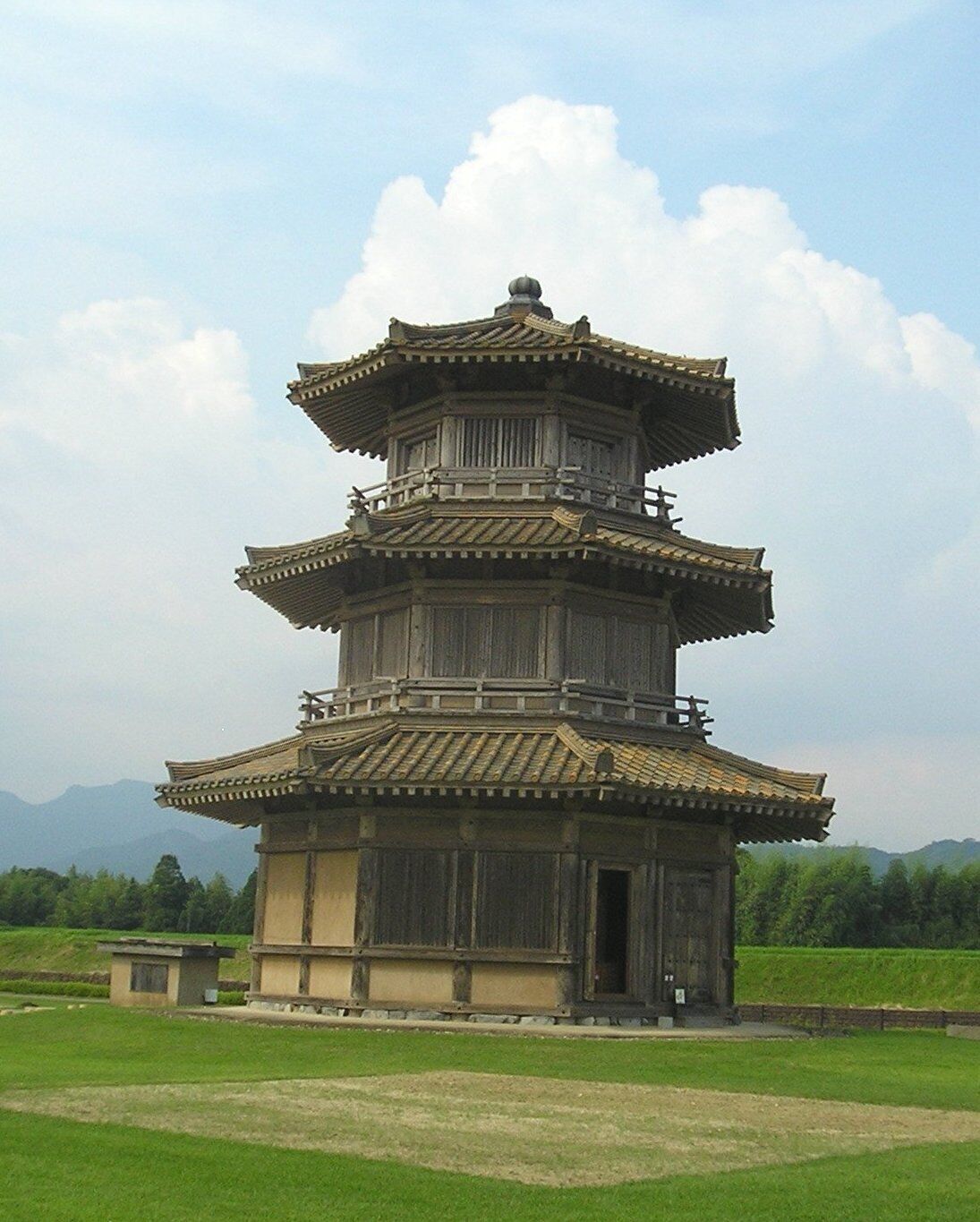 Una pagoda tradicional de varios niveles con techo de tejas, rodeada por un campo cubierto de césped y montañas al fondo.