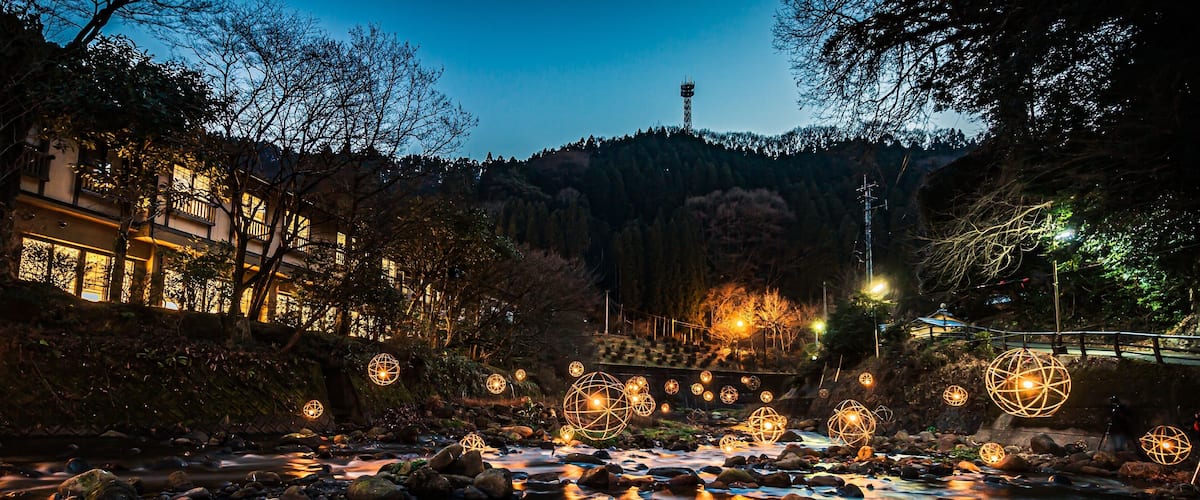A river with rocks and illuminated spherical lights, surrounded by trees and buildings.
