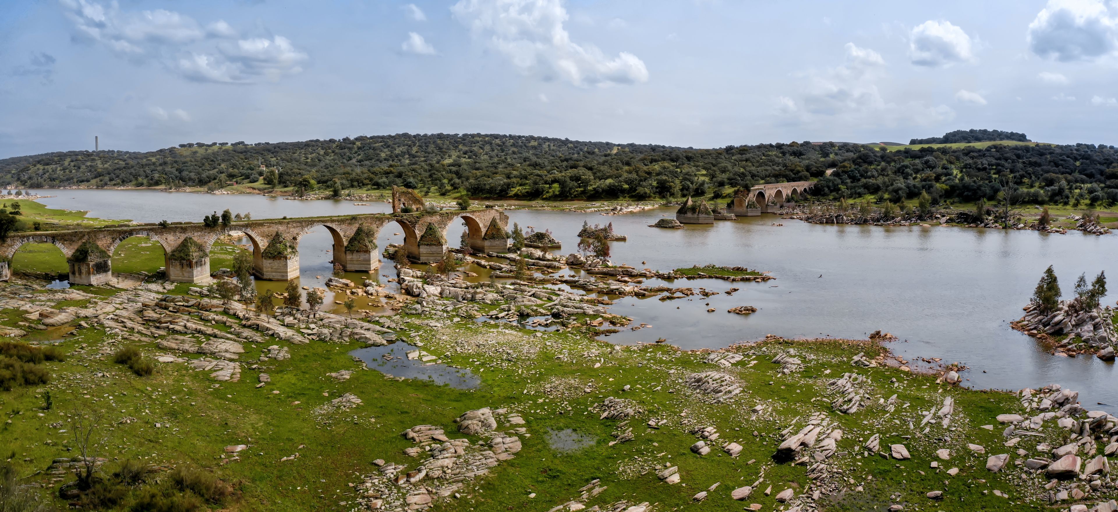 A river with a stone bridge, green grass, and a forested hill in the background.