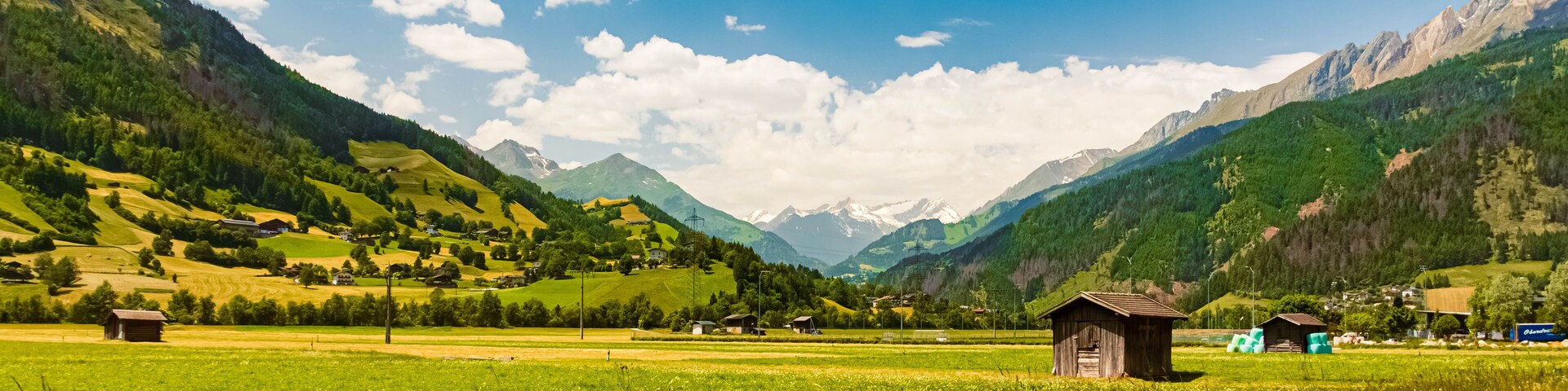 Un paisaje montañoso con campos verdes, casas dispersas y un cielo azul despejado.