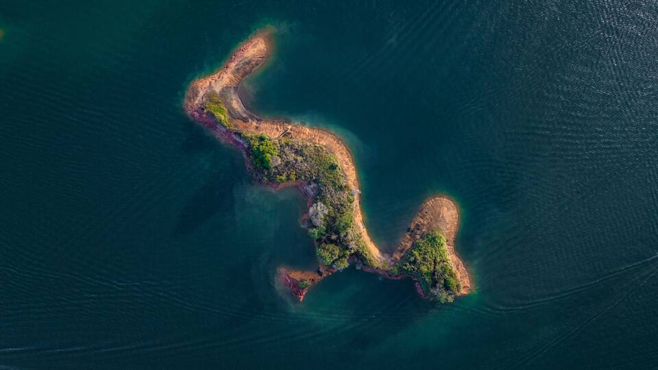 An aerial view of a narrow, elongated island with a mix of green vegetation and sandy areas, surrounded by deep blue ocean waters.