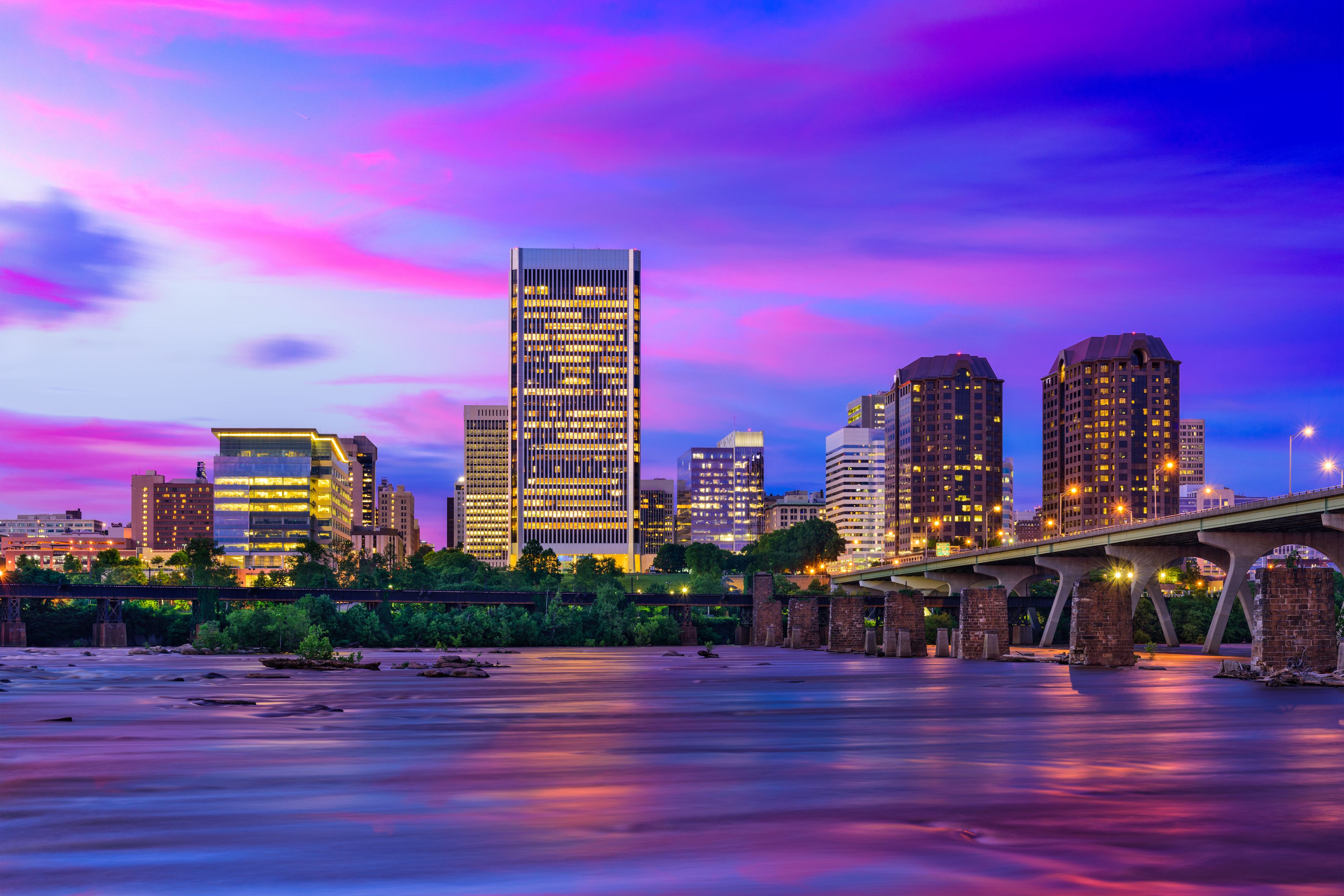 A city skyline at dusk with illuminated buildings and a bridge over a river.