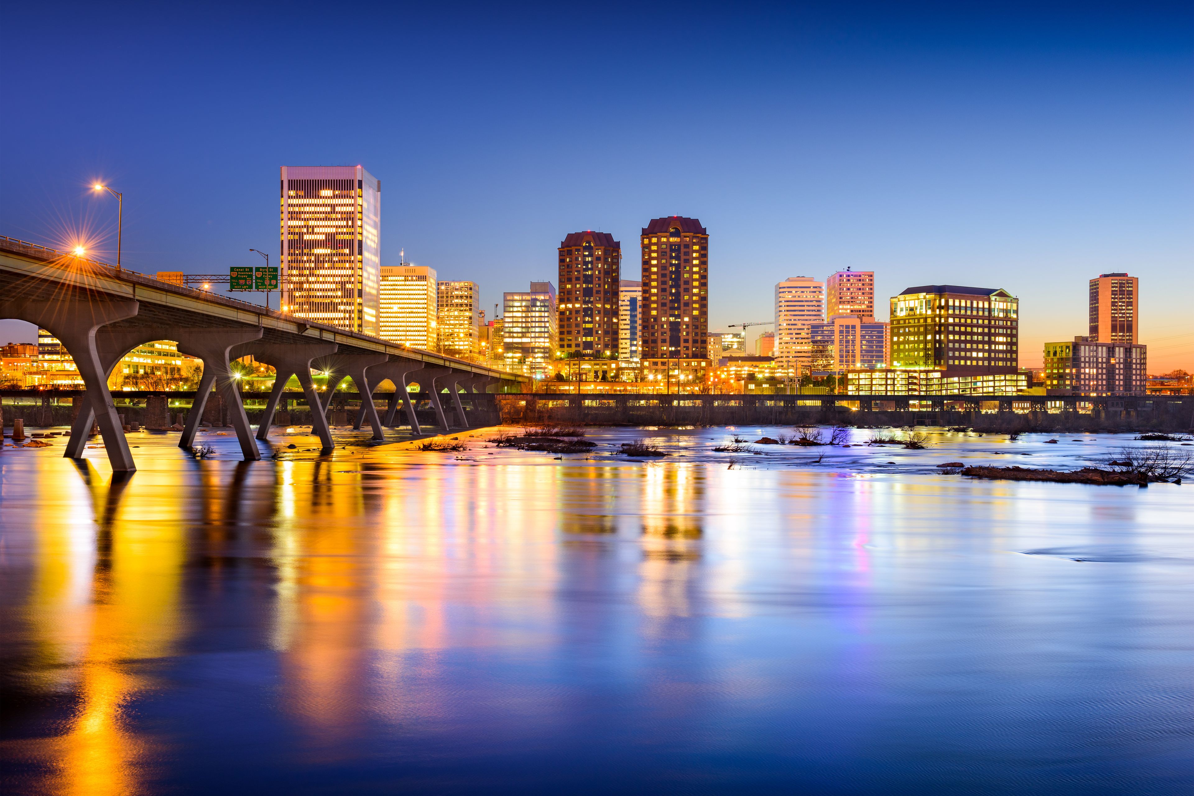 A city skyline at dusk with illuminated buildings and a bridge over a river.