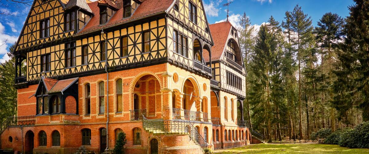 A large, historic half-timbered house with a steep roof, surrounded by a forest.